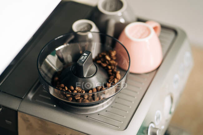 From above of roasted coffee beans in electric grinder against ceramic cups on rack of modern espresso maker at home — Stock Photo