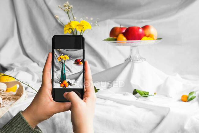 Crop anonymous person taking photo of fresh kumquats and blooming flower bouquet in vase on cellphone on white background — Photo de stock