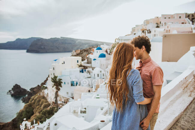 Молодая романтическая пара нежно сближается, стоя на прибрежной улице с белыми типичными домами возле синего волнистого моря в Санторини — стоковое фото