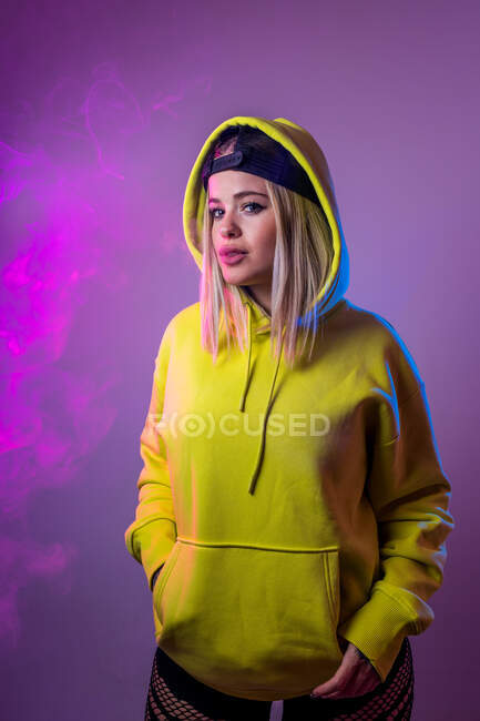 Confident female hipster in hoodie smoking e cigarette in studio on pink background and looking at camera — Stock Photo