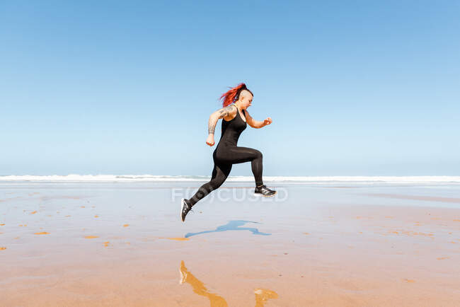 Vista lateral da jogger fêmea ativa com tatuagens pulando na costa do oceano, refletindo na água durante o treinamento — Fotografia de Stock