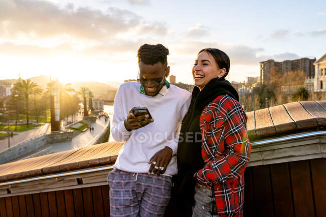 Feliz casal multirracial em vestuário elegante com telefone celular falando na cidade sob céu nublado no crepúsculo — Fotografia de Stock