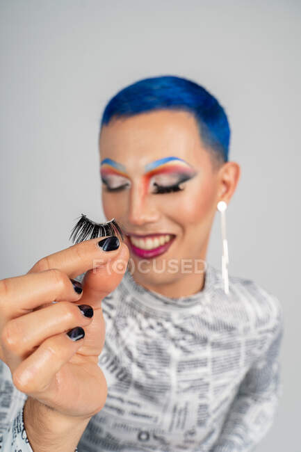 Cheerful drag queen with extravagant bright makeup demonstrating false eyelashes while standing with eyes closed against white background — Photo de stock