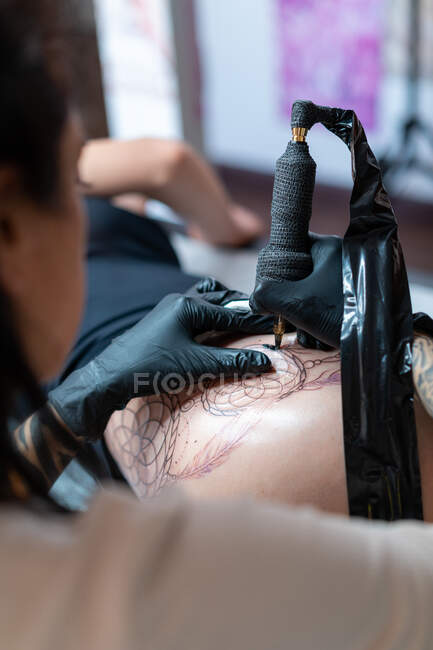 Tatuaje femenino con tatuaje de dibujo de máquina en el cuerpo del cliente irreconocible en el salón - foto de stock