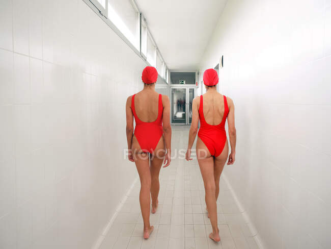 Visão traseira de jovens nadadoras anônimas em trajes de banho vermelhos andando em corredor estreito — Fotografia de Stock