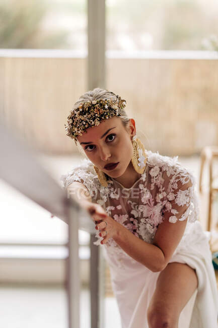 Joven hembra en elegante vestido de novia blanco bohemio y botas de tacón alto con corona ornamental y pendientes de pie en la escalera y mirando a la cámara - foto de stock