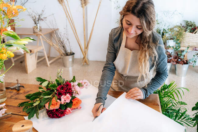 Зверху урожаю щаслива етнічна молода жінка-квітка в фартусі посміхається і ріже папір для обгортання, а також організовує елегантний букет зі свіжих різнокольорових квітів — стокове фото