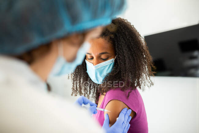 Especialista médica feminina irreconhecível em uniforme de proteção, luvas de látex e máscara facial vacinando paciente afro-americana na clínica durante o surto de coronavírus — Fotografia de Stock