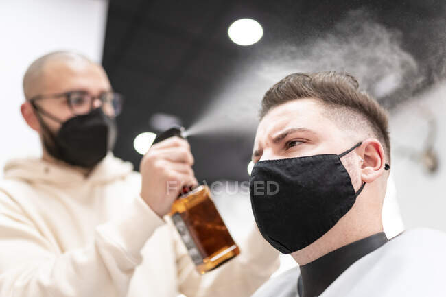 Male stylist in textile mask spraying haircut of client with liquid from bottle in barbershop — Photo de stock