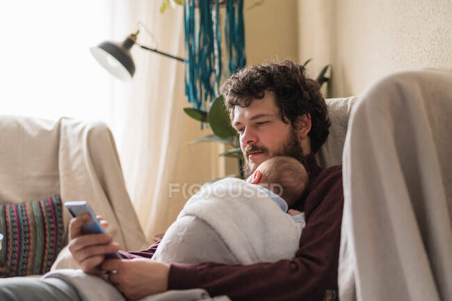 Papà adulto che abbraccia il bambino anonimo mentre naviga in internet sul cellulare in casa durante il giorno — Foto stock