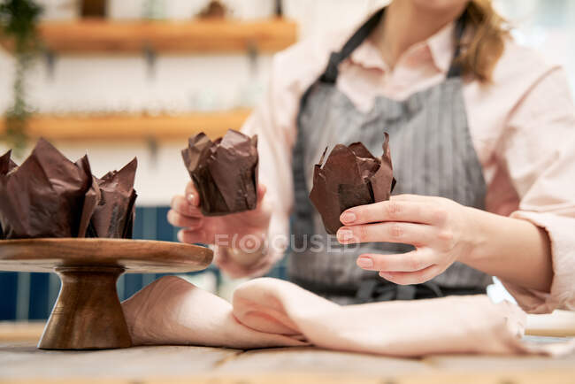 Colheita fêmea irreconhecível em avental mostrando sobremesa assada em forros de papel na cozinha da casa — Fotografia de Stock