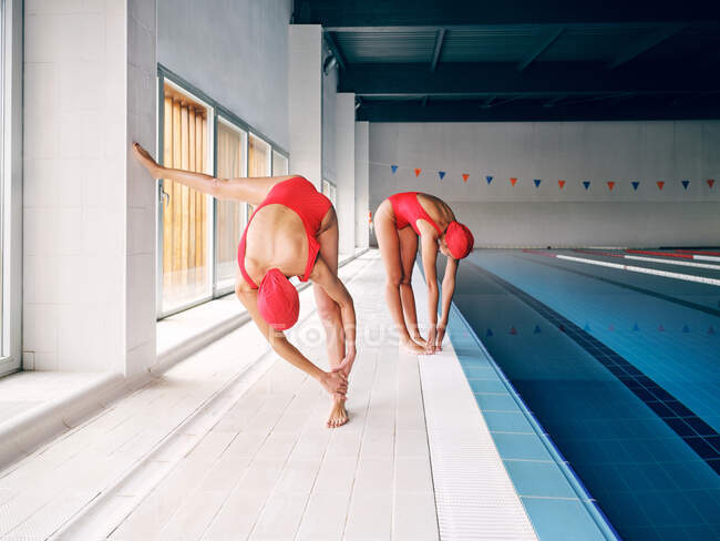 Deportivas anónimas en traje de baño rojo estirando las piernas mientras realizan doblado hacia adelante contra la piscina a la luz del día - foto de stock
