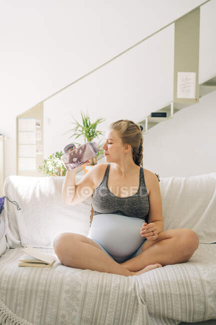 Jovem grávida em roupas esportivas com garrafa de água sentada com pernas cruzadas no sofá em casa — Fotografia de Stock