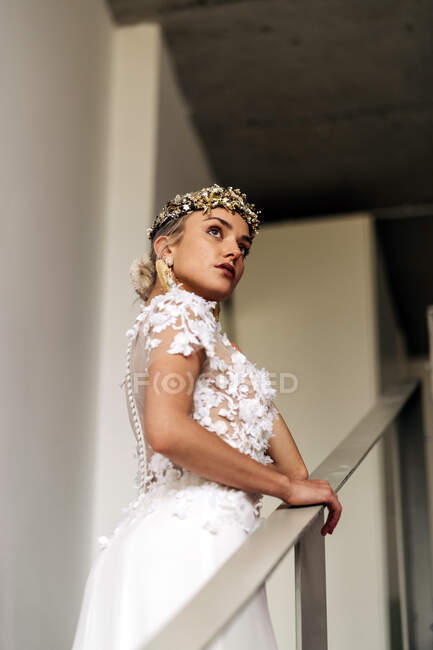 Side view of young female in stylish bohemian white bridal dress and high heeled boots with ornamental wreath and earrings standing on stairway and looking away — Foto stock
