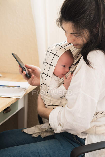 Side view of mom with cute little child in carrier text messaging on cellphone in house room — Stock Photo