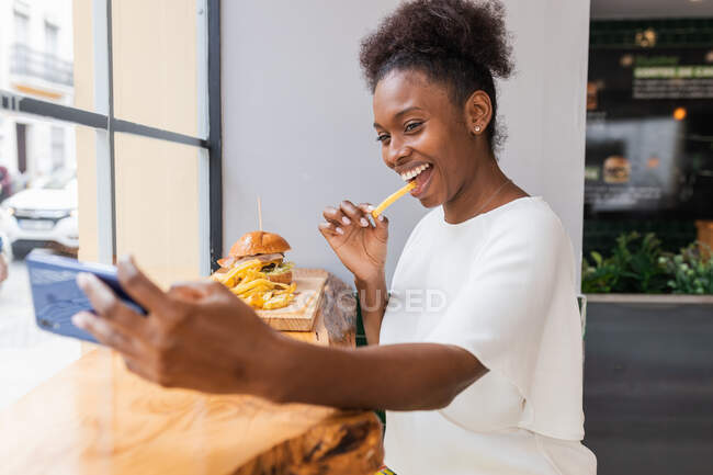 Vista lateral alegre joven afroamericana femenina en blusa blanca disfrutando de deliciosas papas fritas y hamburguesas mientras toma selfie en el teléfono inteligente en el restaurante de comida rápida - foto de stock