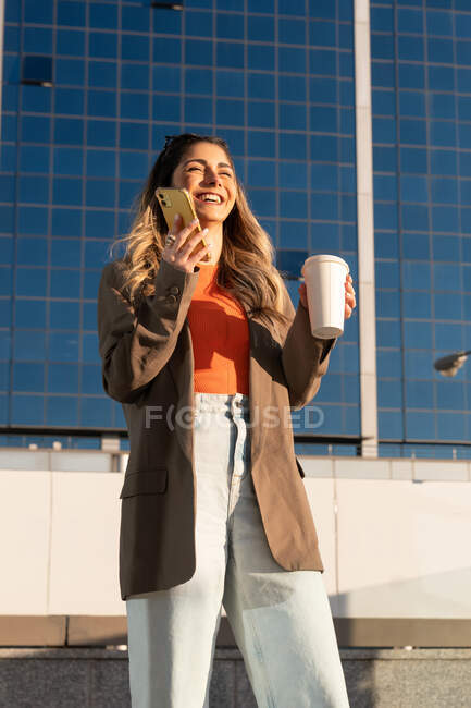 Joven mujer alegre en ropa elegante con bebida caliente para llevar y teléfono celular mirando hacia adelante en la ciudad - foto de stock