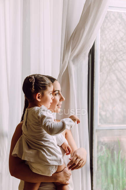 Vista lateral de la madre étnica con linda hija pequeña mirando por la ventana mientras está de pie en la habitación en casa - foto de stock
