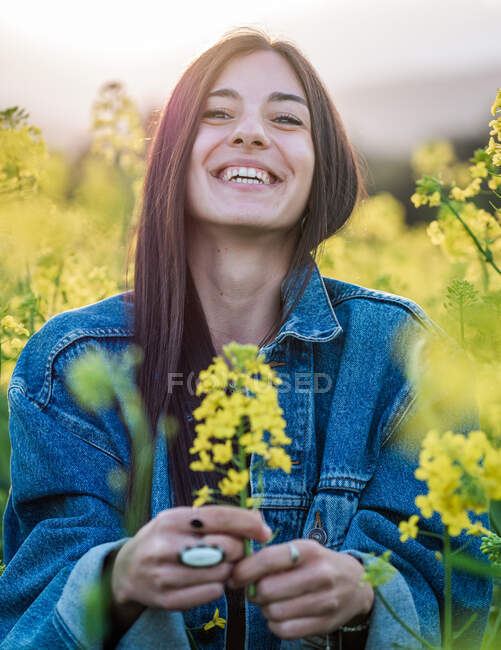 Delighted young brunette in denim jacket laughing merrily demonstrating fragrant yellow rapeseed flowers on hands standing on blooming field on sunny day — Stock Photo