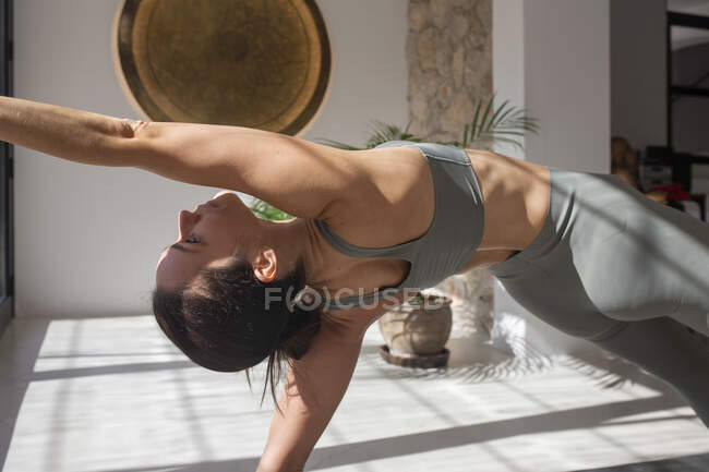 Vista lateral de la hembra en ropa deportiva preparándose para la pose Camatkarasana mientras practica yoga en casa - foto de stock