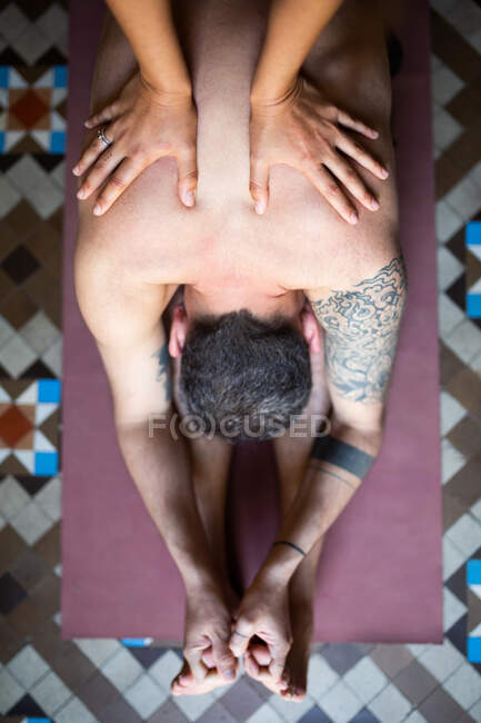 Visão superior do homem flexível anônimo fazendo ioga em Pascimottanasana com a ajuda do instrutor pessoal da colheita enquanto sentado no tapete em casa — Fotografia de Stock