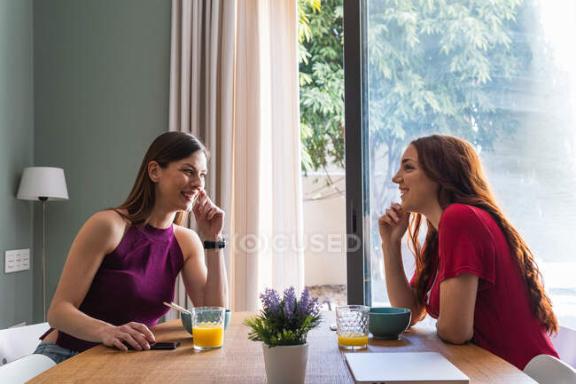 Fröhliche junge Freundinnen in lässigen Outfits beim gemeinsamen leckeren Mittagessen und Smartphone-Gebrauch, während sie am Esstisch in einer modernen Wohnung sitzen und plaudern — Stockfoto