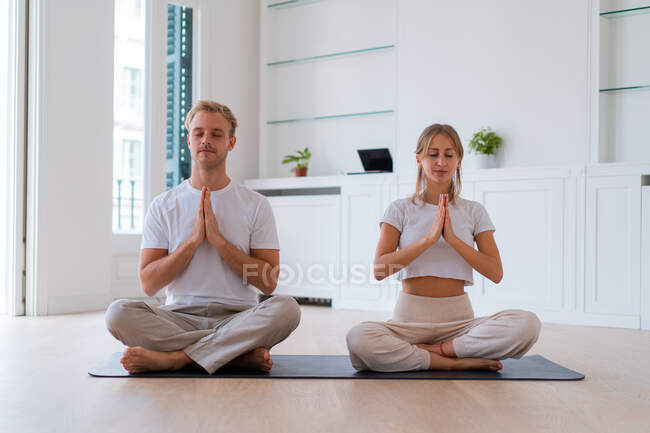 Мирная пара сидит в позе лотоса с молитвенными руками, практикуя йогу вместе и медитируя с закрытыми глазами — стоковое фото
