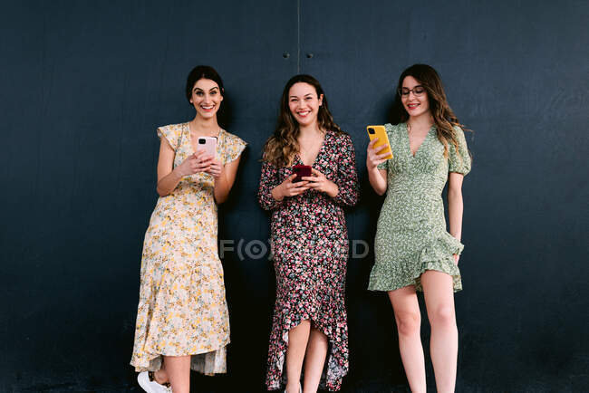 Content young best female friends in trendy apparel with cellphones standing on urban walkway against wall — Stock Photo