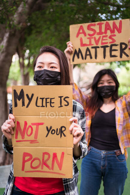 Mujeres étnicas con máscaras sosteniendo carteles protestando contra el racismo en la calle de la ciudad y mirando a la cámara - foto de stock