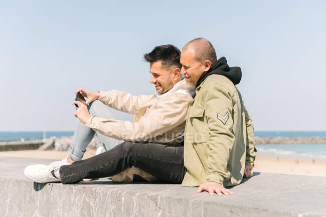 Вид сбоку романтическая молодая пара геев в модных нарядах улыбается и делает селфи, отдыхая на каменной скамейке возле песчаного пляжа под безоблачным голубым небом — стоковое фото