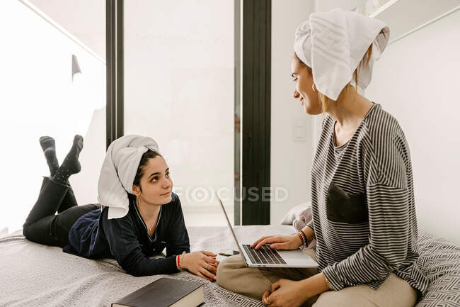 Happy young ethnic lady in casual clothes and towel on head lying on bed and looking at girlfriend working remotely on laptop at home — Stock Photo