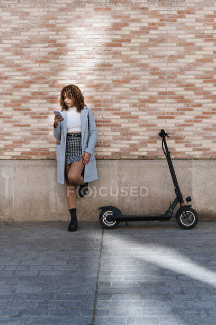 Young African American female with curly hair wearing blue coat browsing on mobile phone while standing against brick wall and looking at screen with smile — Stock Photo