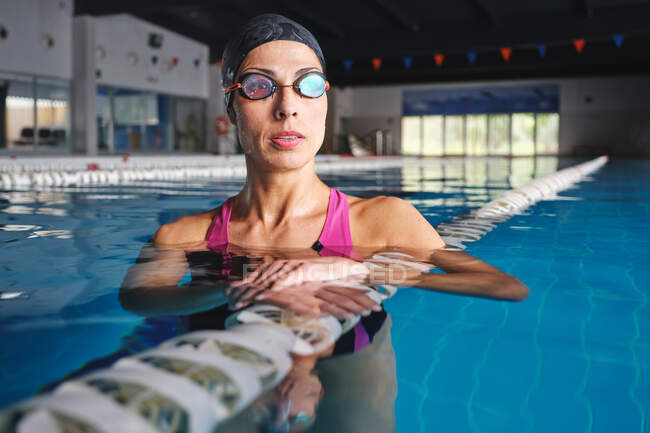 Adult sportswoman in goggles and swimwear leaning on lane line in swimming pool with transparent water and looking away — Stock Photo