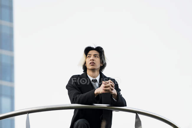 Young ethnic male entrepreneur in coat leaning on fence while looking away against contemporary city house — Stock Photo