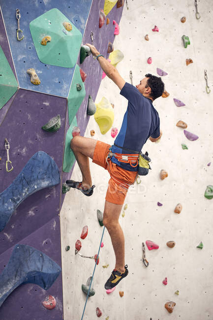 Vista lateral macho montañero en arnés escalada pared artificial en el centro de bouldering - foto de stock