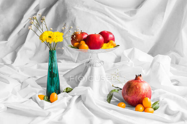 Schmackhafter Granatapfel mit roten Äpfeln und Kumquats in der Nähe der Vase mit blühenden Chrysanthemen auf weißem Knitterstoff — Stockfoto