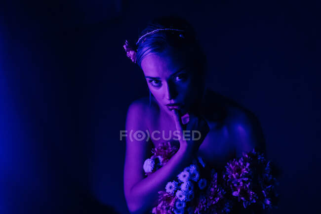 Attraktive junge barbusige Frau in weißen Hosen mit einem Strauß frischer bunter Blumen sitzt auf Hockern gegen grelles Neonlicht im dunklen Studio — Stockfoto