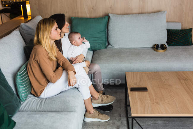 Vista lateral de alto ángulo de contenido par de mujeres lesbianas sentadas en el sofá con el bebé lindo y viendo la película juntos en la sala de estar - foto de stock