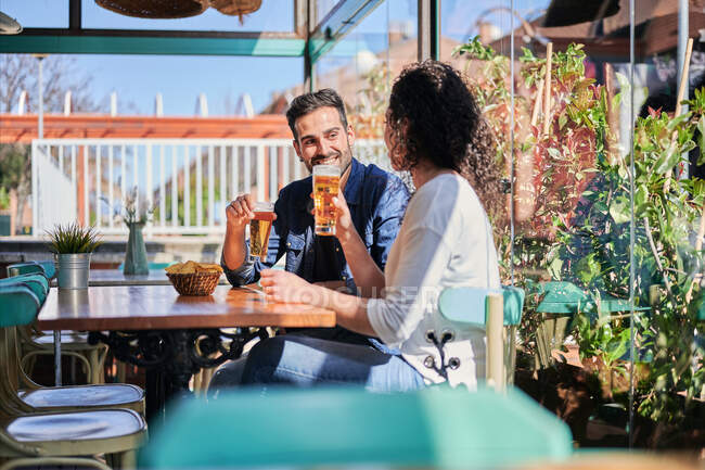 Fröhliches ethnisches Paar mit Gläsern Bier und Kartoffelchips im Gespräch, während es sich im Sonnenlicht anschaut — Stockfoto
