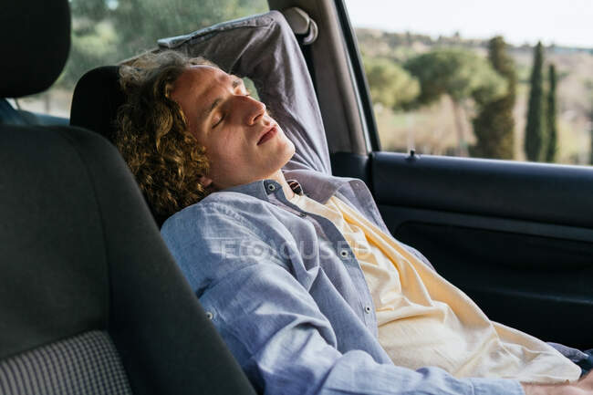 Розслаблений молодий мандрівник спить на водійському сидінні сучасного автомобіля, відпочиваючи під час поїздки по сільській місцевості в літній день — стокове фото