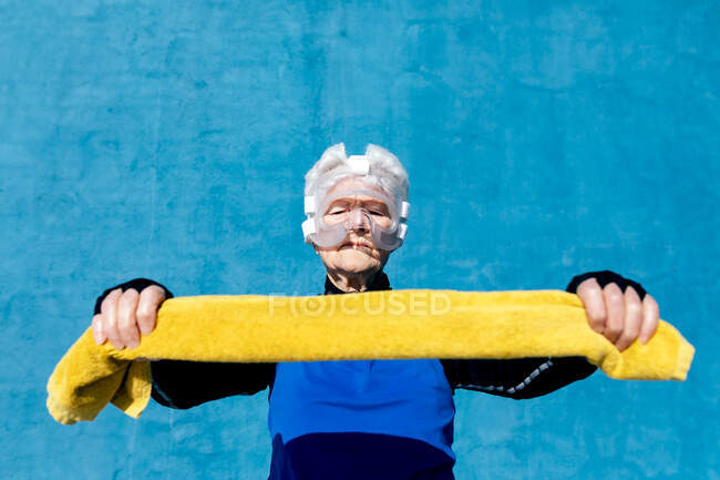 Serious mature female in activewear in protective boxing head guard with towel in the hands standing on blue background with closed eyes — Stock Photo