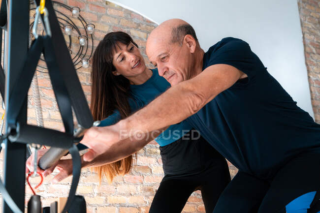 Улыбающаяся инструктор помогает мужчине делать упражнения на пилатес реформатор во время тренировки в тренажерном зале — стоковое фото