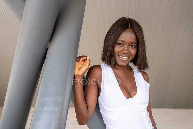Happy attractive African American female wearing stylish white dress with low neckline standing near concrete poles on street and looking at camera — Stock Photo