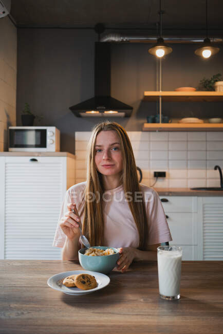 Jeune femme avec cuillère et bol profitant de savoureux anneaux de maïs tout en regardant la caméra dans la cuisine — Photo de stock