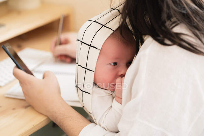 Cropped unrecognizable mom with cute little child in carrier text messaging on cellphone in house room — Stock Photo