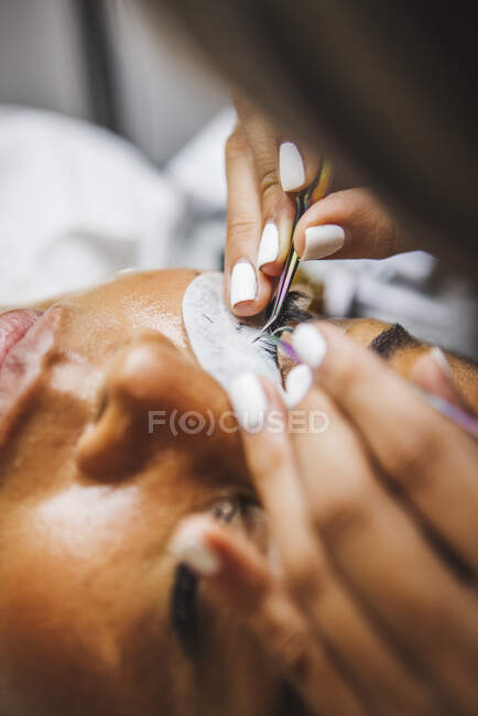 Hoher Winkel der Ernte unkenntlich Kosmetiker mit Pinzette Anwendung gefälschte Wimpern für die Verlängerung auf das Auge des ethnischen Kunden im Salon — Stockfoto