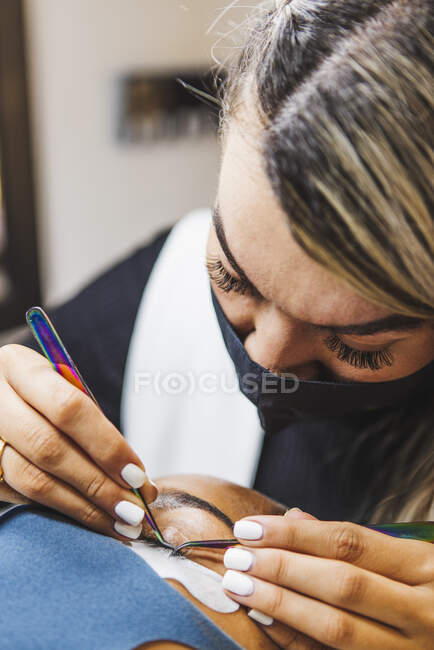 Cosmetologist with tweezers applying fake eyelashes for extension on eye of ethnic client with face protective mask in salon during coronavirus pandemic — Fotografia de Stock
