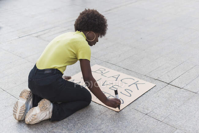 High angle side view of African American female activist writing Black Lives Matter and making placard for protest against racism in city — Foto stock