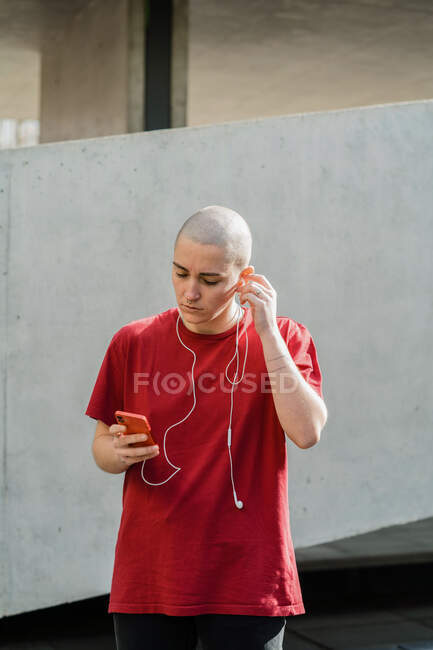 Giovane donna omosessuale in t-shirt e auricolari con cellulare che guarda lo schermo mentre ascolta musica — Foto stock