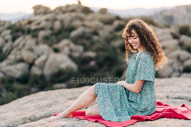 Young barefoot female in blue maxi sundress sitting on red plaid on rough rocky hilltop and looking down — Stock Photo